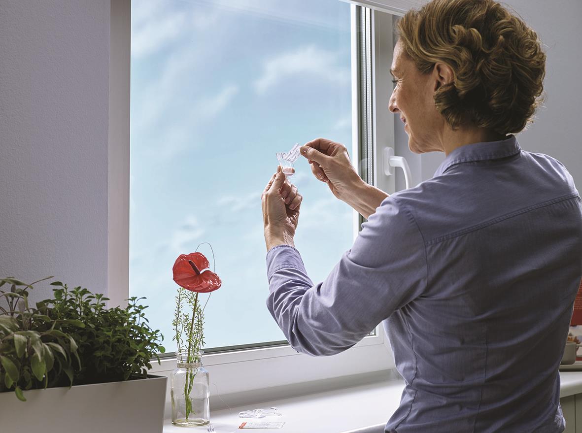 Kvinna sätter upp en självhäftande krok på fönstret med Tesa Powerstrips, inomhus med växter på fönsterbrädan