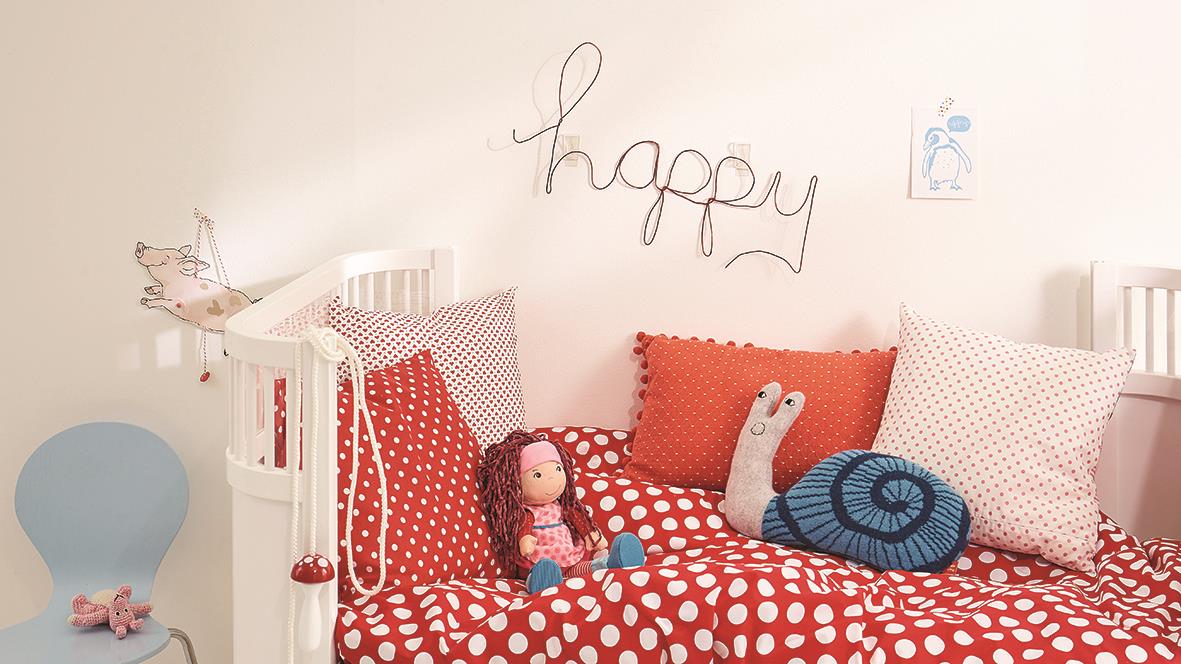 Sovrum med röda och vita sängkläder, leksaker och väggdekorationer, inklusive ordet happy i trä.