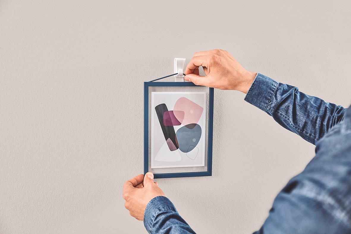 Person hänger upp en konstnärlig bild i ett vitt rum med självhäftande krok