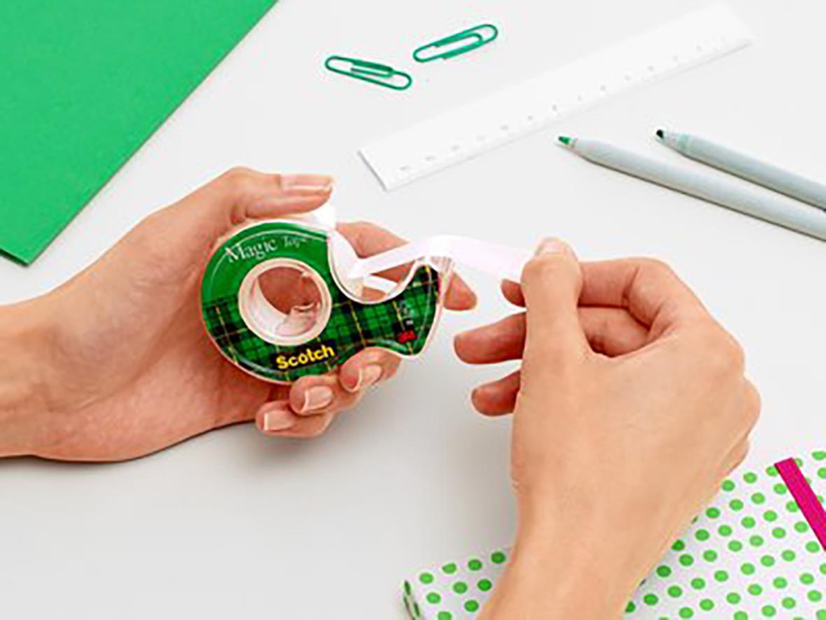 Person håller i Scotch Magic tejp med hållare, omgiven av papper, gem och penna på ett skrivbord