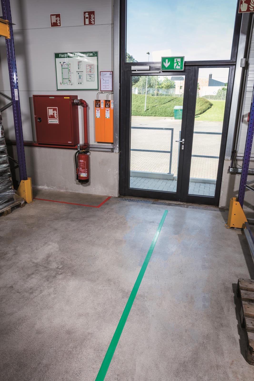 Golvtejp i grönt markerar en linje på betonggolv i ett industriutrymme nära en dörr med nödutgångsskylt