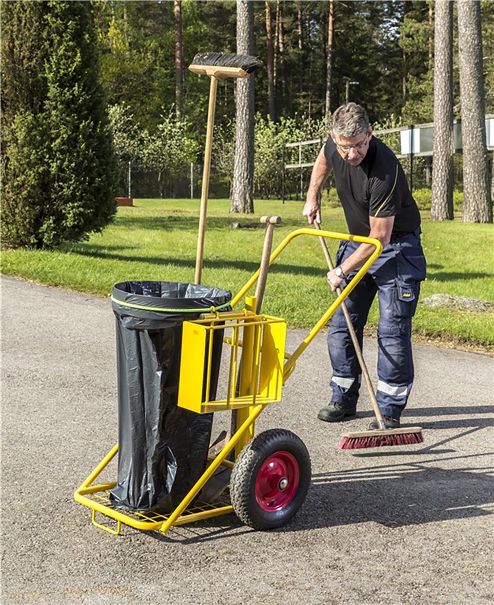 Person städar utomhus med gul städkärra och sopborste på en grusväg omgiven av träd och gräs