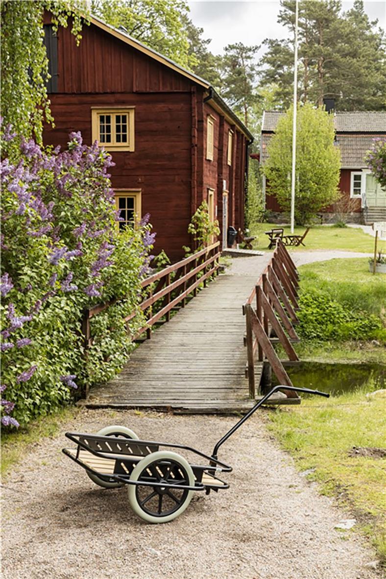Träflak dragkärra med hjul och handtag framför en liten träbro i en trädgård