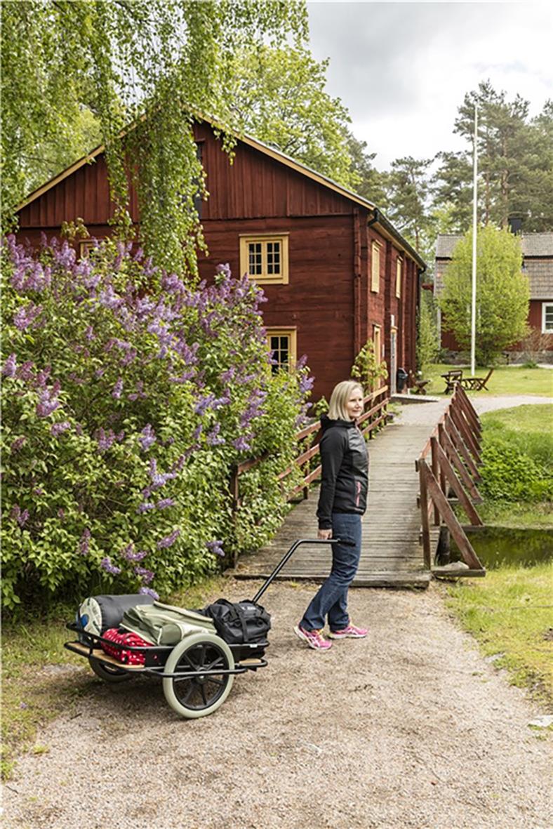Kvinna med dragkärra med träflak framför ett rött hus och gröna träd