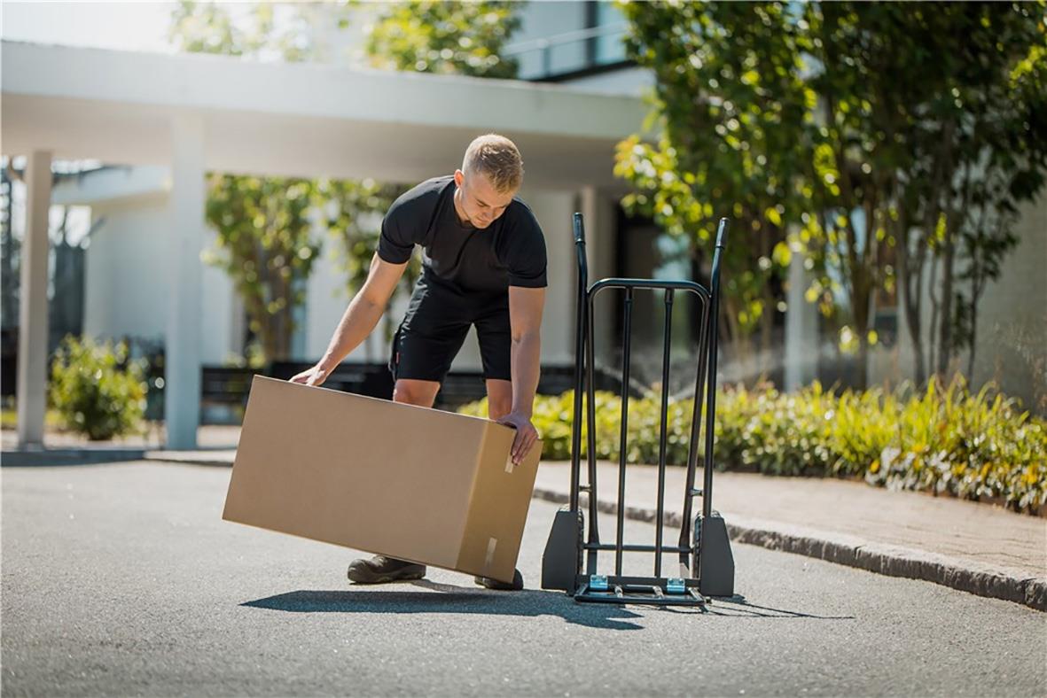 Man flyttar en stor kartong utomhus med en handkärra bredvid