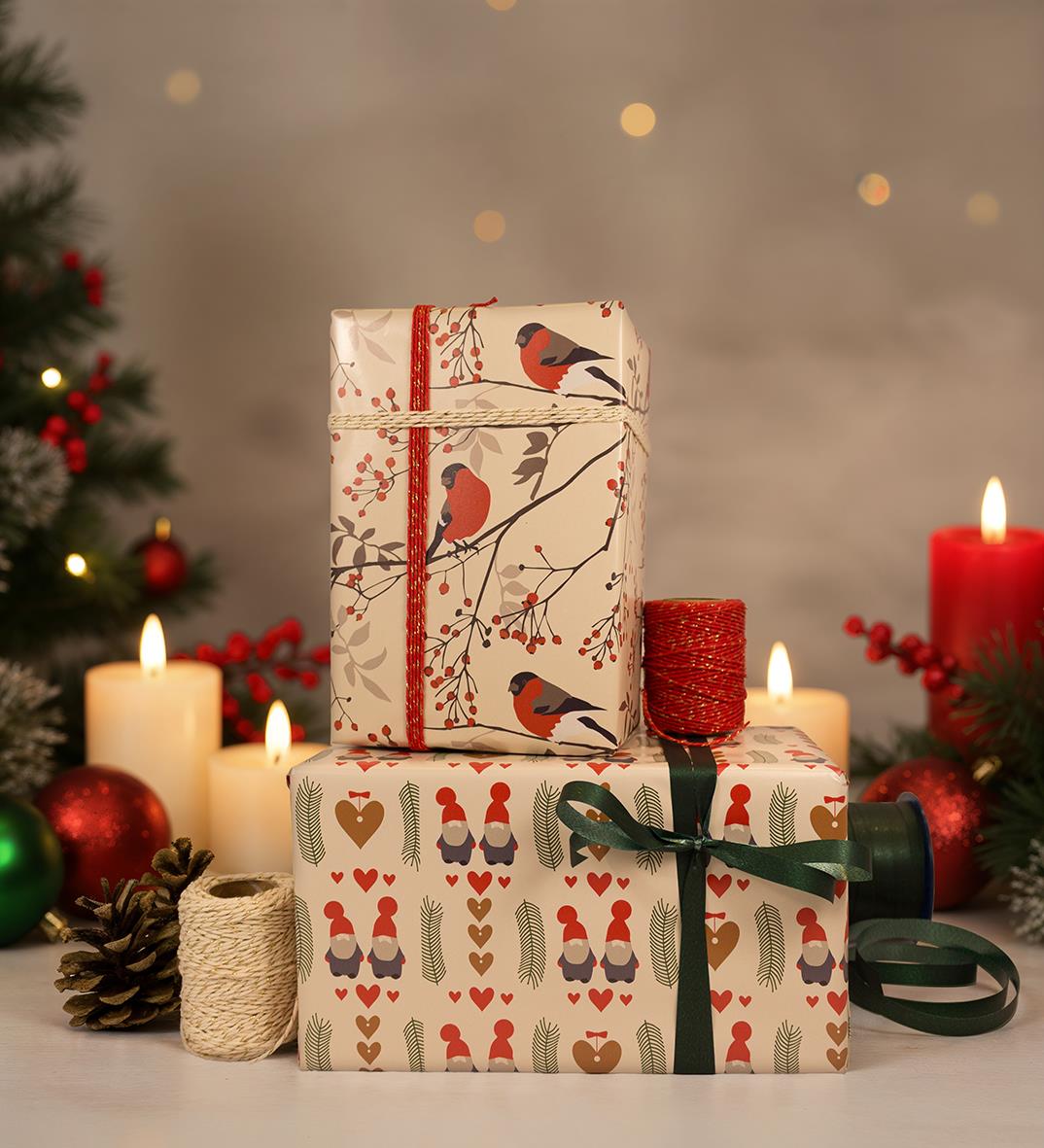 Julklappar inslagna i julpapper med domherrar och tomtemotiv, dekorerade med gröna band och omgivna av levande ljus