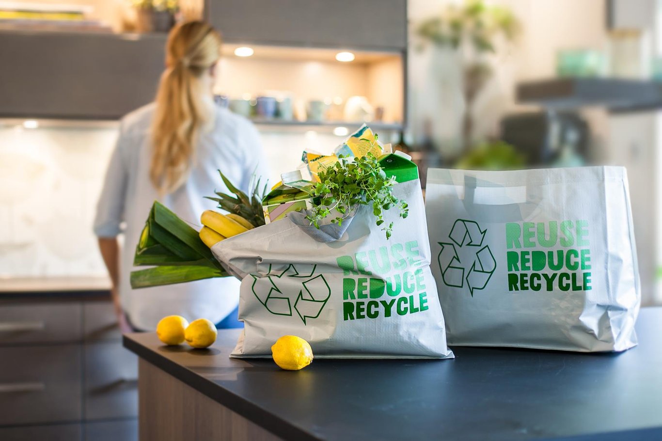 Två återvinningspåsar med texten Reduce Recycle på köksbänk, fyllda med grönsaker och frukt, i ett modernt kök