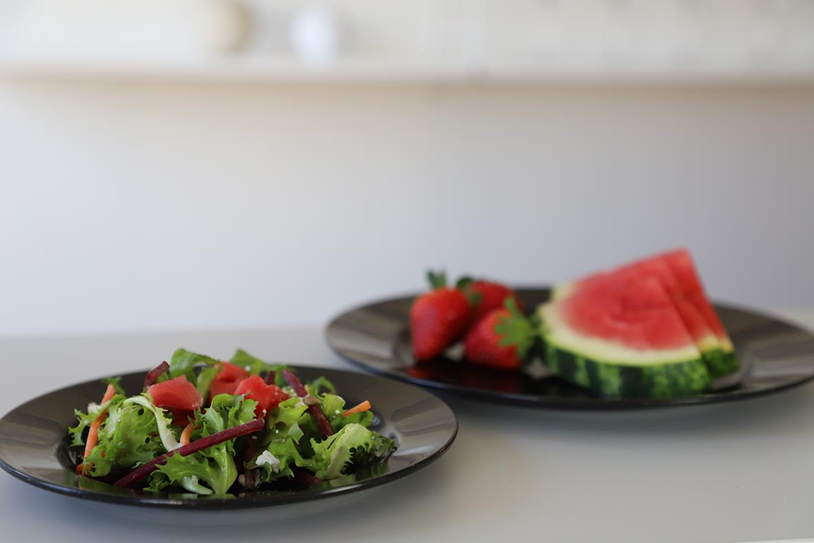 Svart tallrik med sallad och en annan tallrik med vattenmelon på en vit yta