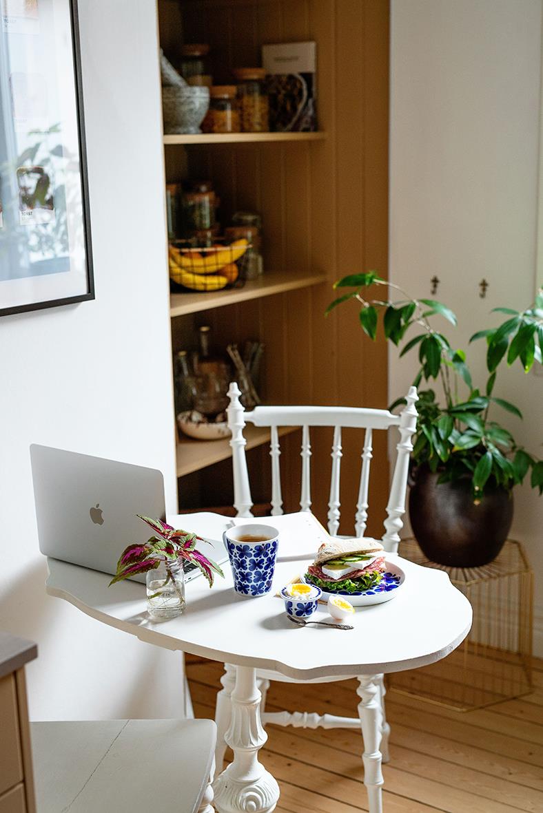 Mysig köks- eller vardagsrumsscen med ett runt vitt bord, en vit stol, en växt i stor kruka och ett öppet skåp med burkar och frukt i bakgrunden