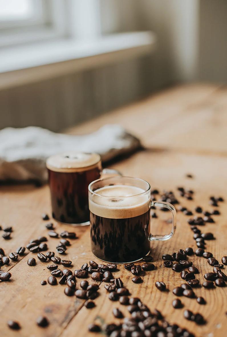 Två glas med mörk kaffe på träbord omgivna av kaffebönor, bakgrund av fönster och köksmiljö