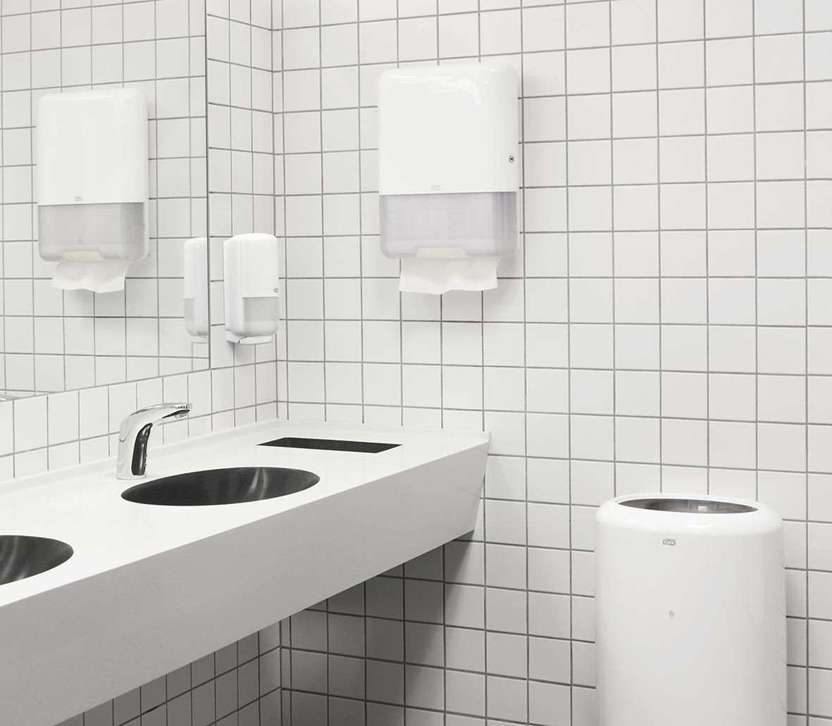 Två pappershandduksautomater och en papperskorg i ett badrum med vita kaklade väggar