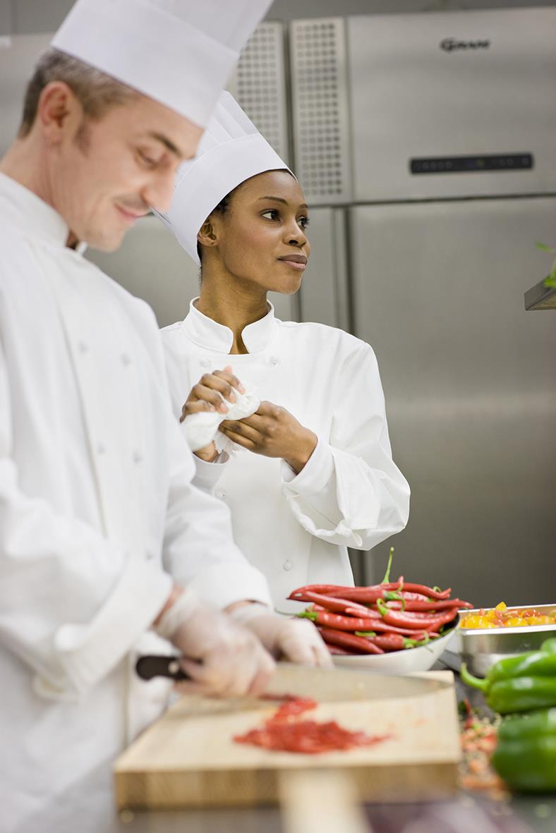 Två kockar i kök, en skär kött och den andra håller en pappershandduk, köksutrustning och grönsaker syns i bakgrunden