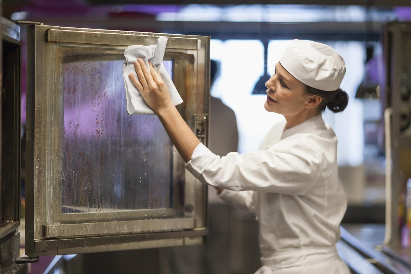 Kvinna i arbetskläder torkar en fönsterruta i ett kök eller köksliknande miljö