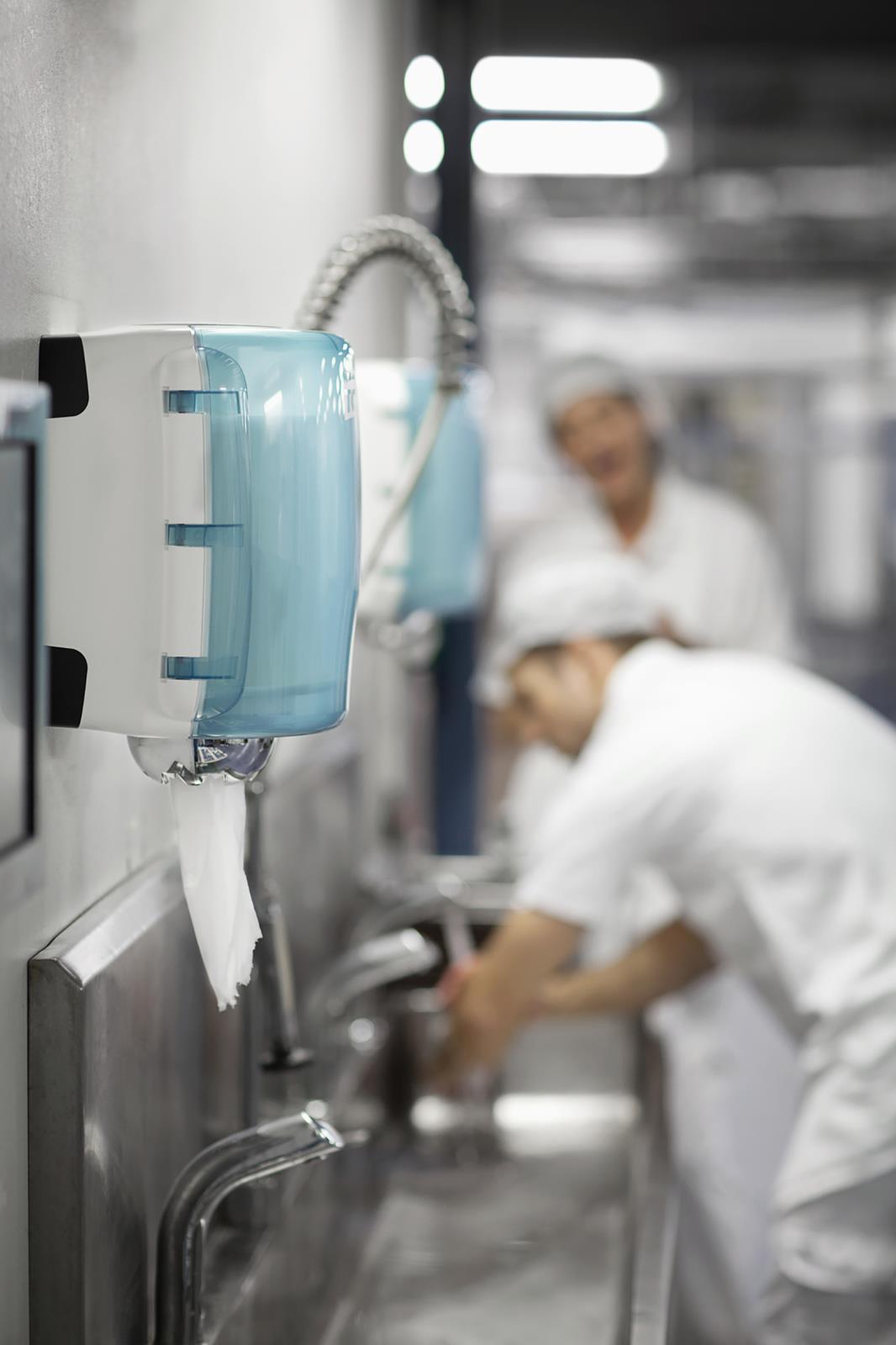 Två personer i arbetskläder tvättar händer i ett industriellt kök eller tvättrum med en blå handduksdispenser på väggen