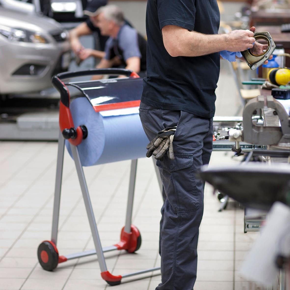 Person som använder en blå industrirulle Tork W1/2 E-Kraft 3-lg på en arbetsplats med verktyg och andra personer i bakgrunden