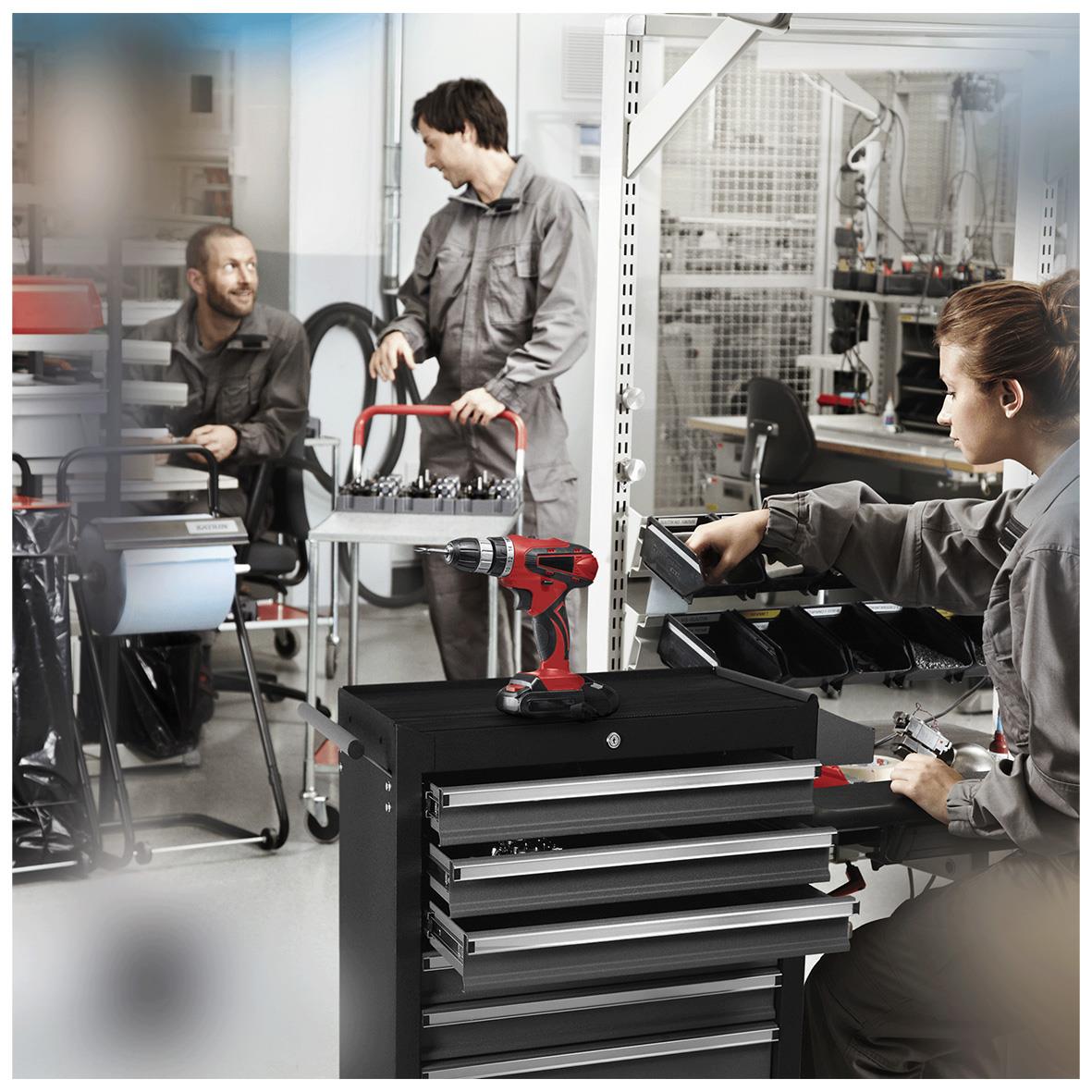 Två personer i ett industrilabb arbetar med verktyg och maskiner, en person sitter vid ett skrivbord och en annan står med ett verktyg