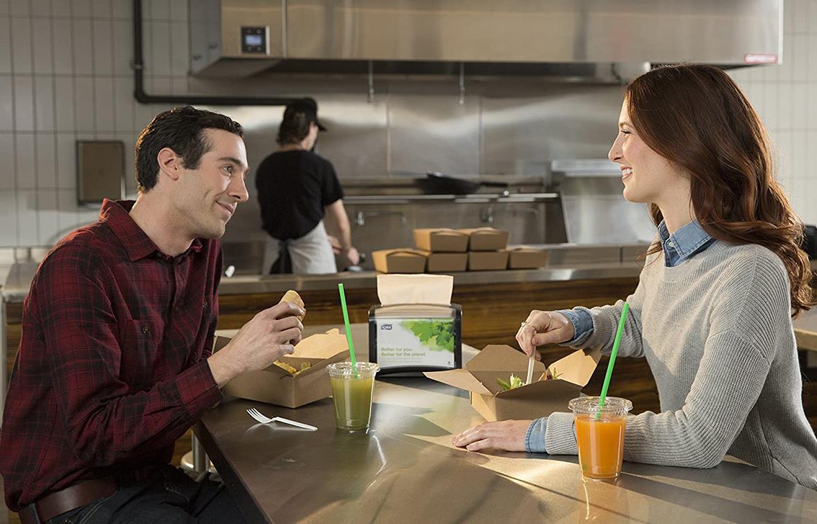 Två personer som pratar och dricker drycker på ett café eller restaurang, med en kök i bakgrunden.