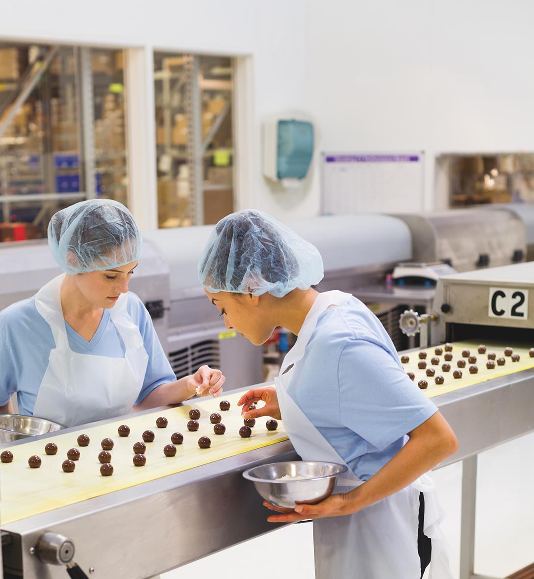 Två personer i arbetskläder och hårskydd arbetar med chokladpraliner på en fabriksgolvet
