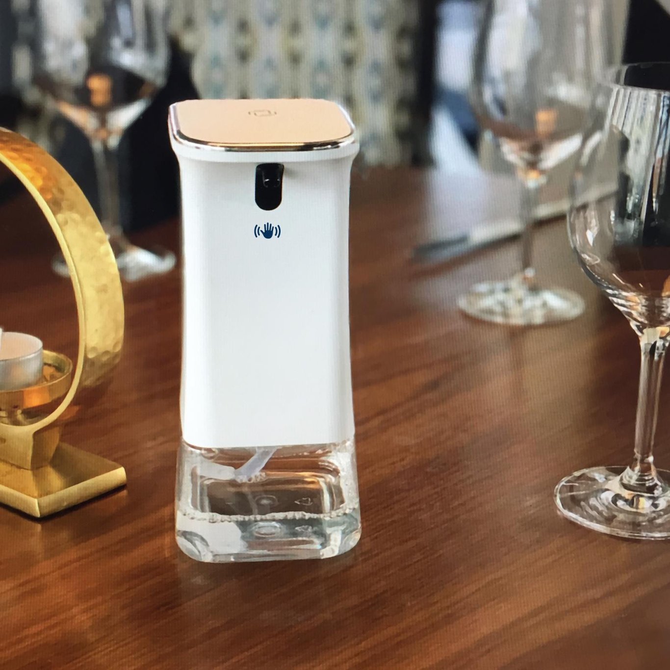 Automatisk handdesinfektionsdispenser på bordet med glas och dekorationer i bakgrunden