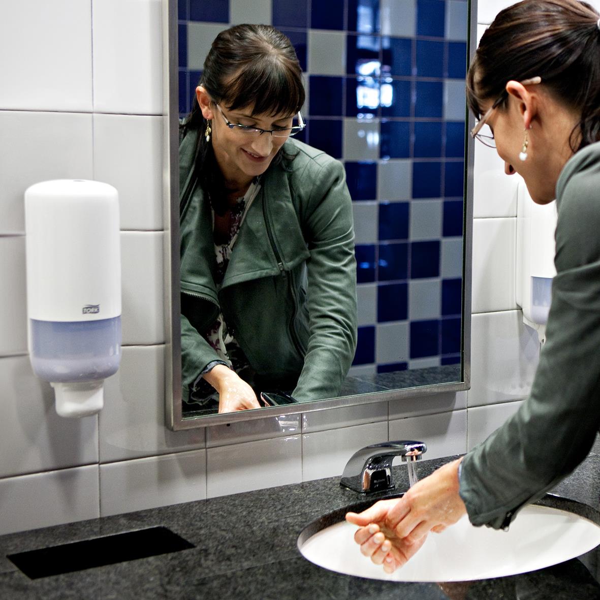 Två kvinnor använder en vit plastdispenser för handdesinfektion i ett badrum med vita och blå kakelväggar