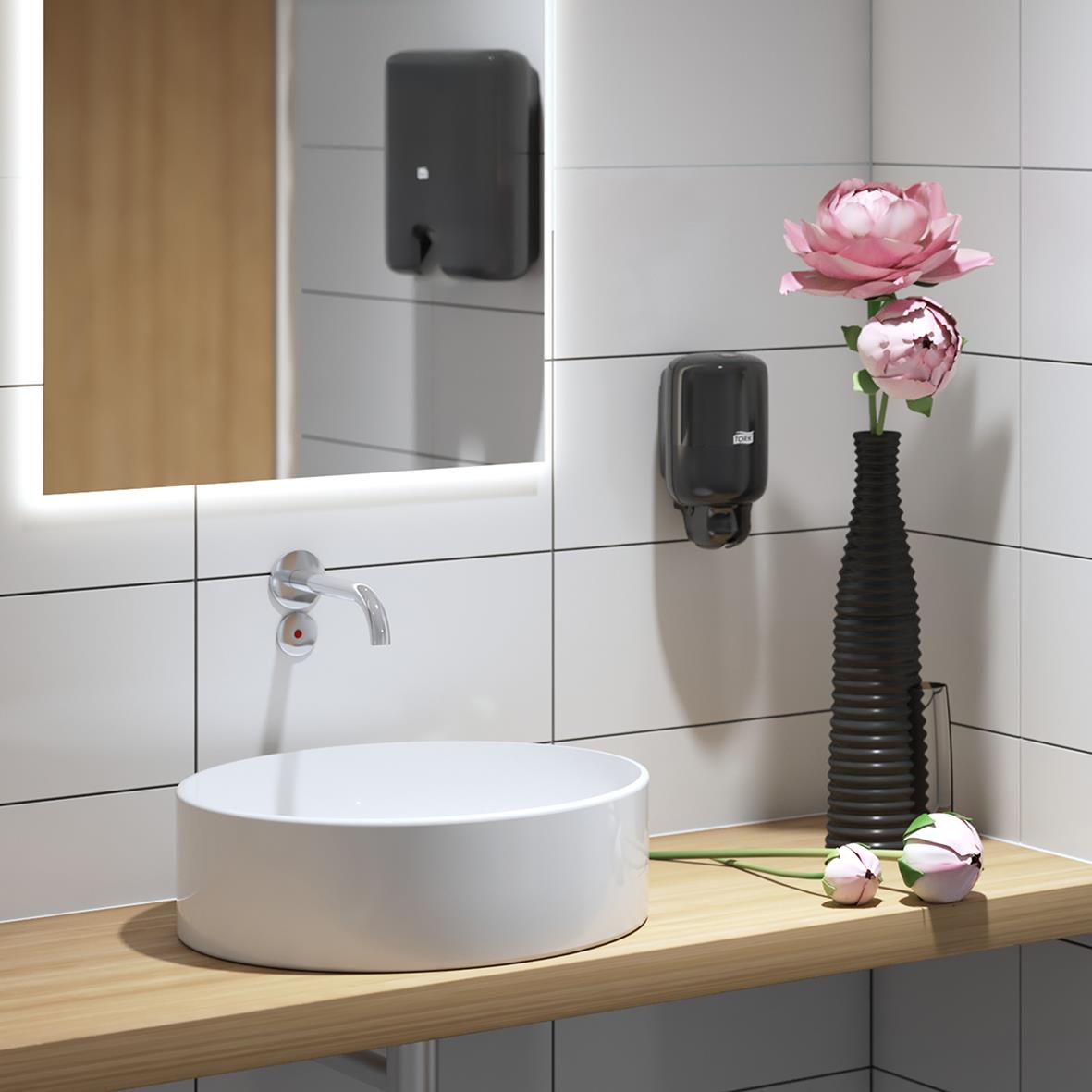Svart Tork S2 Mini plastdispensersystem på vägg ovanför handfat med spegel och blomsterarrangemang