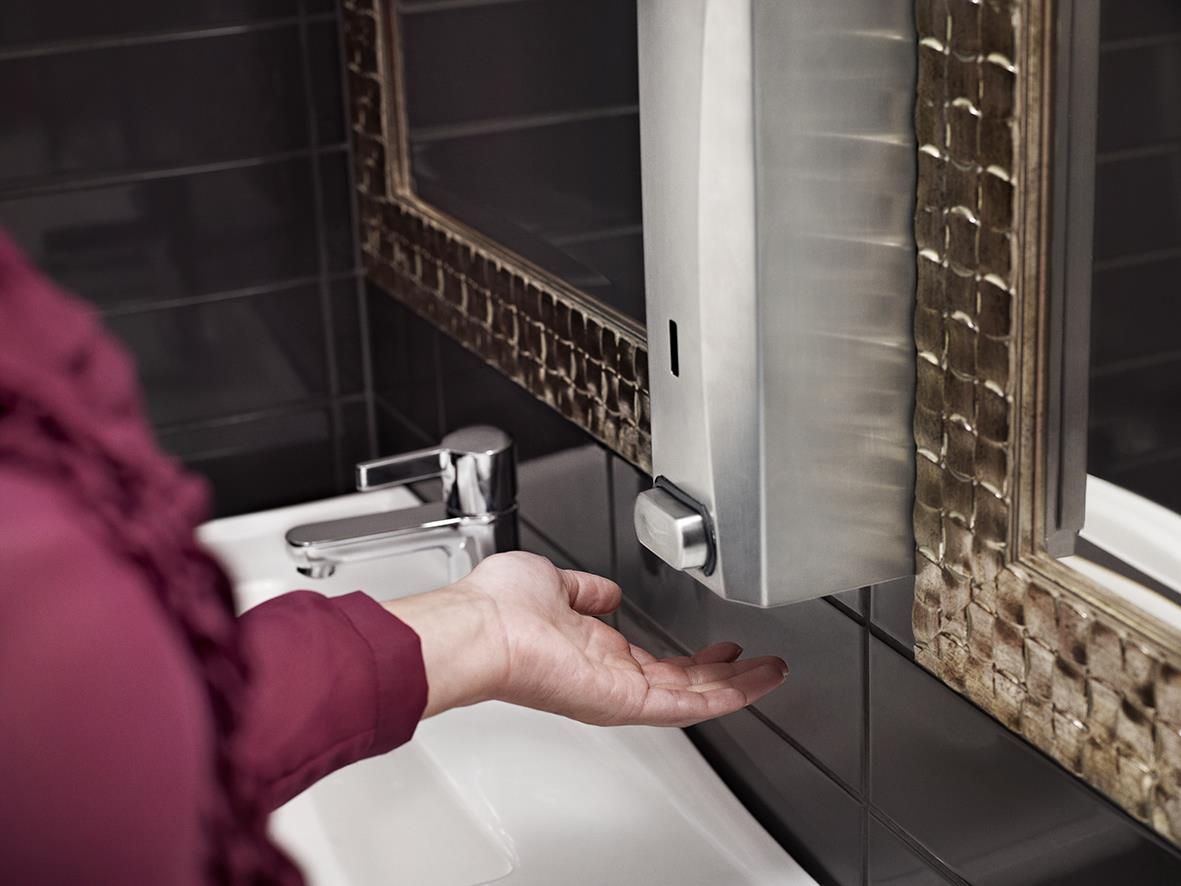 Person som använder Katrin tvåldispensator i badrum med svart kakel och spegel