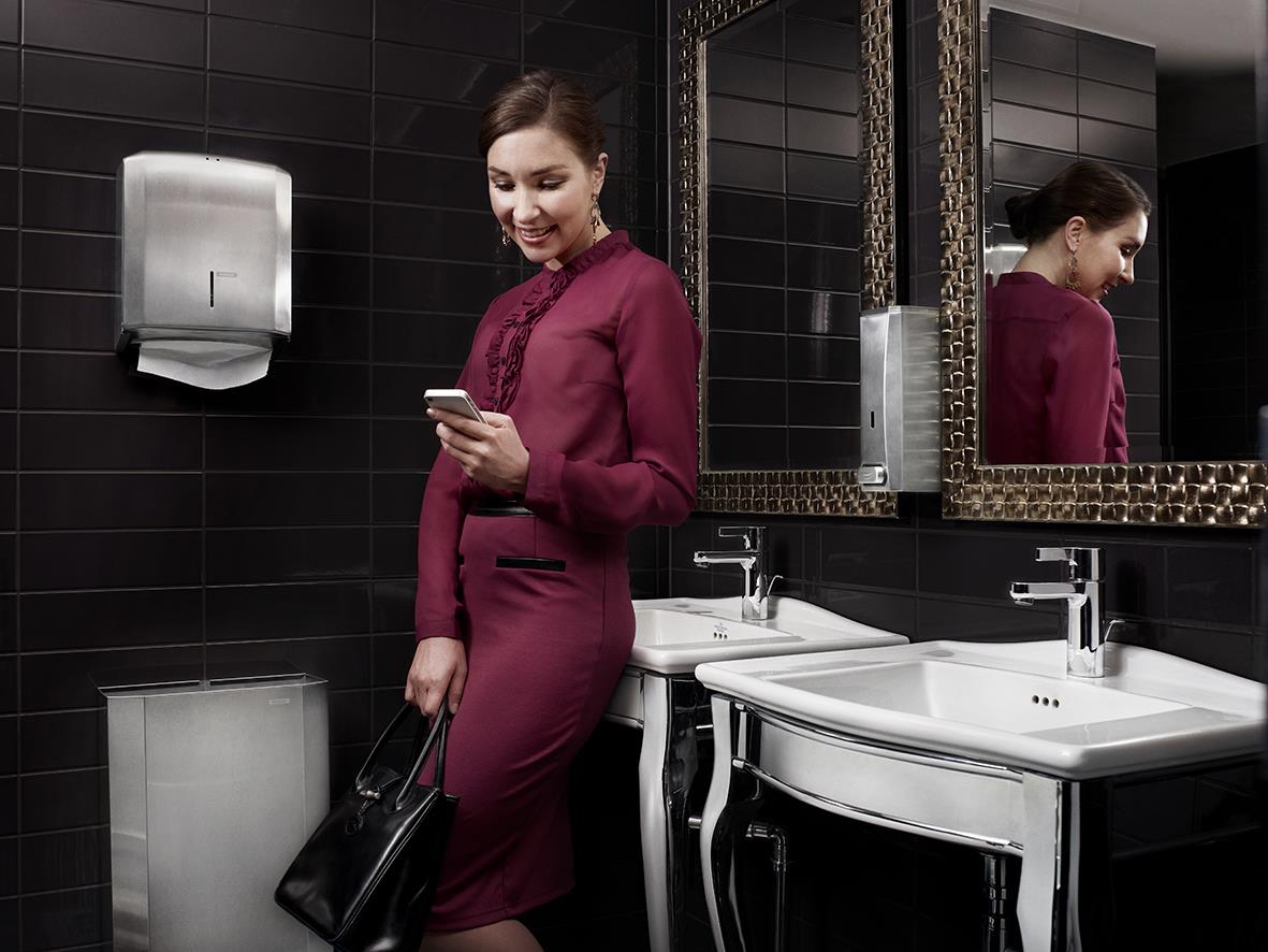 Kvinna i mörkröd klänning använder Katrin tvål dispenser i badrum med svart kakel och speglar