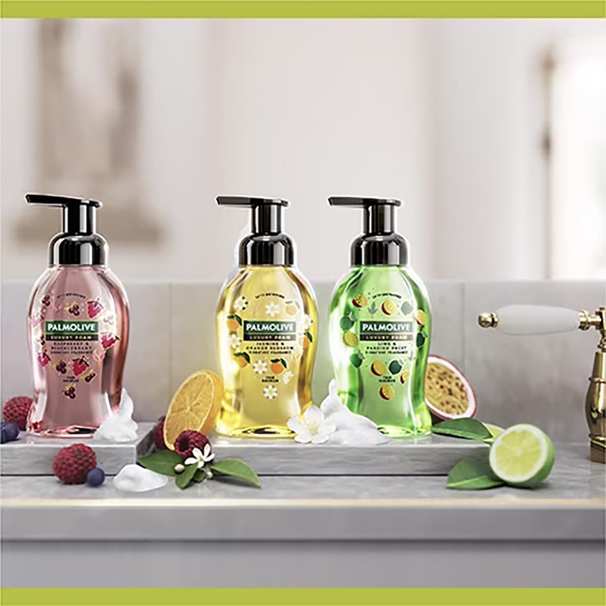 Tre Palmolive Luxury Foam skumtvålar i olika färger med citrusfrukter och blommor runt om på en köksbänk