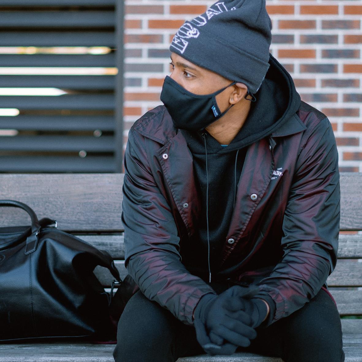 Ung man i svart munskydd, mössa och jacka sitter på en parkbänk utomhus