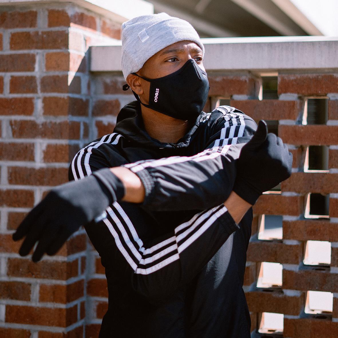 Person i svart jacka och vit mössa bär svart munskydd utomhus vid en tegelvägg