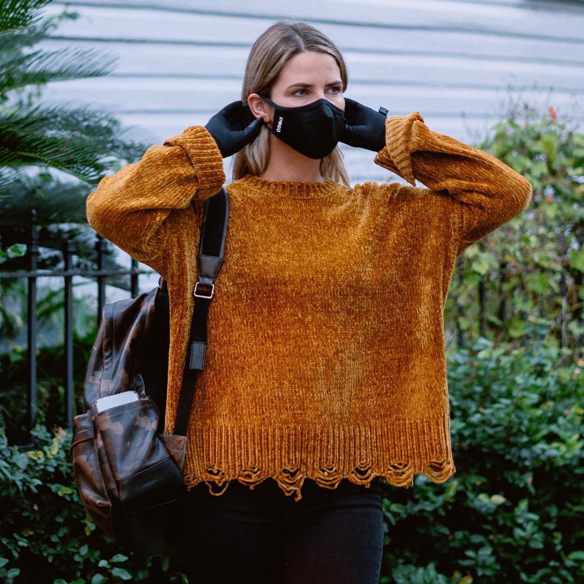 Kvinna bär svart munskydd och orange tröja utomhus med växter och staket i bakgrunden