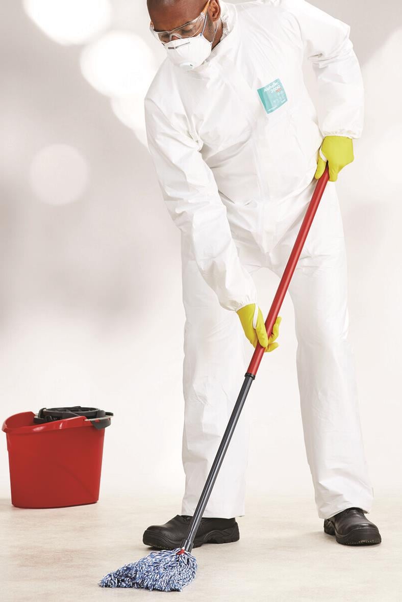 Person i skyddskläder och mask som moppar golvet med en gul flersgångshandske på handen