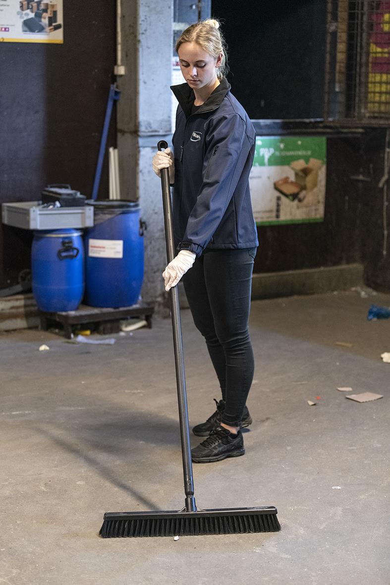 Kvinna som städar golv med en lång svart sopborste i en industriell miljö, bär jacka och handskar
