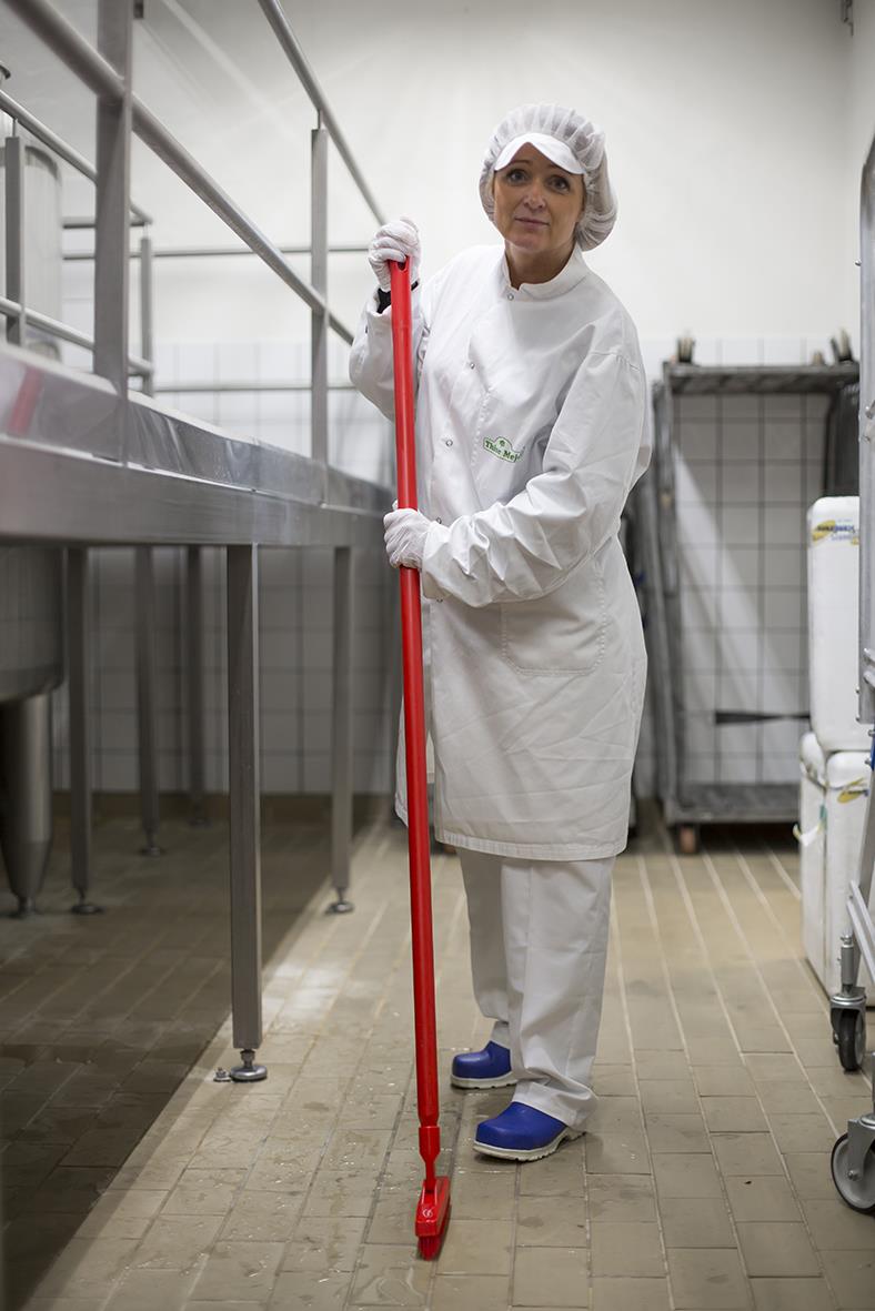 Person i skyddskläder med hårmössa och handskar som håller en blå extra hård fogborste i ett kök eller industriell miljö