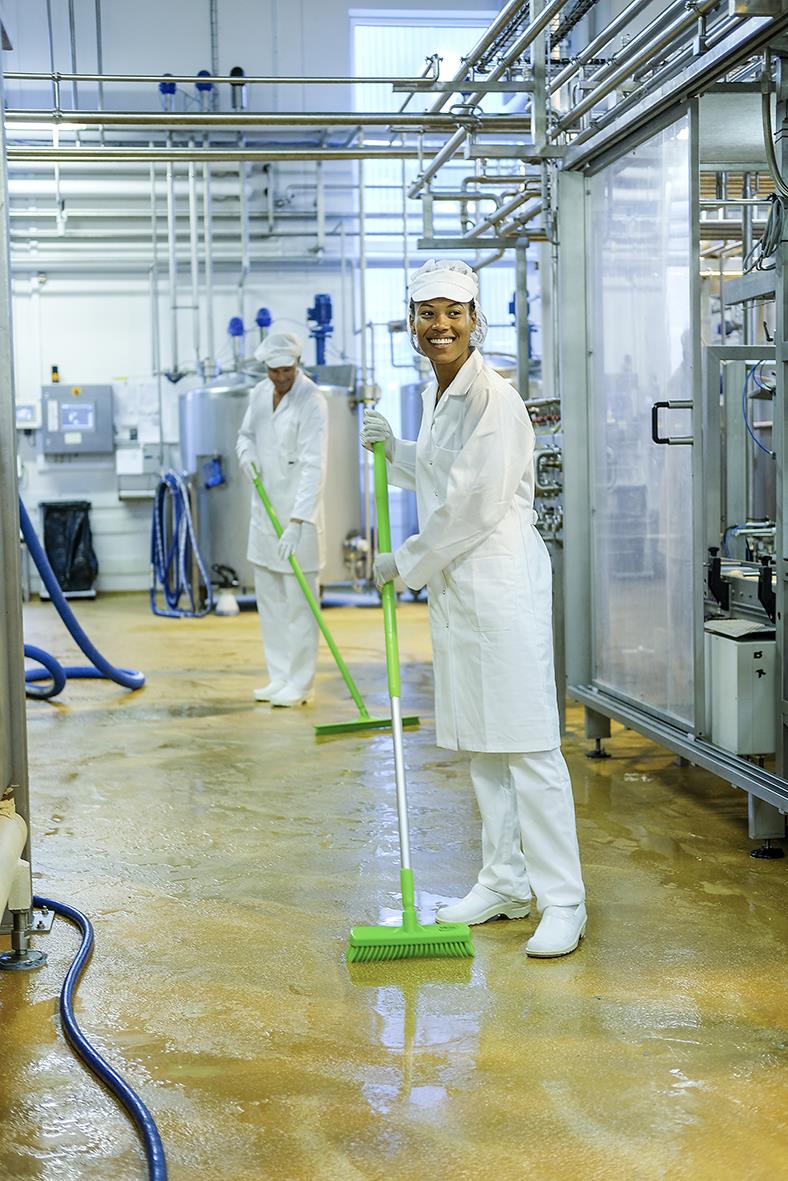 Två personer i vita arbetskläder som städar i ett industriellt kök eller laboratorium med gröna städredskap