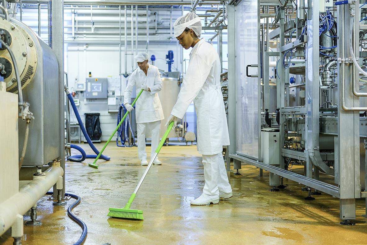 Två personer i skyddskläder städar ett industriellt golv med gröna moppverktyg