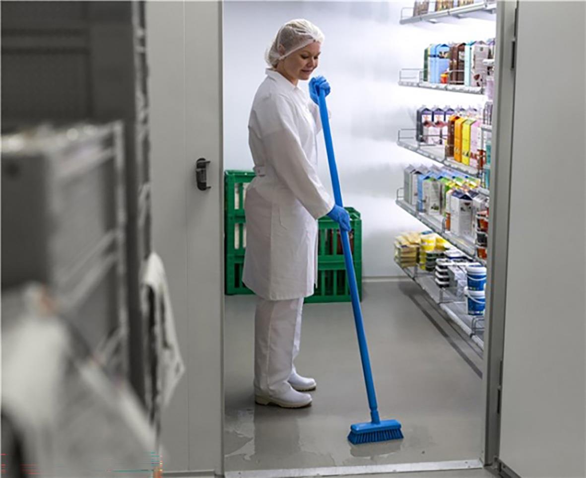 Person i skyddsdräkt som städar golvet i ett laboratorium eller kök med en blå borste