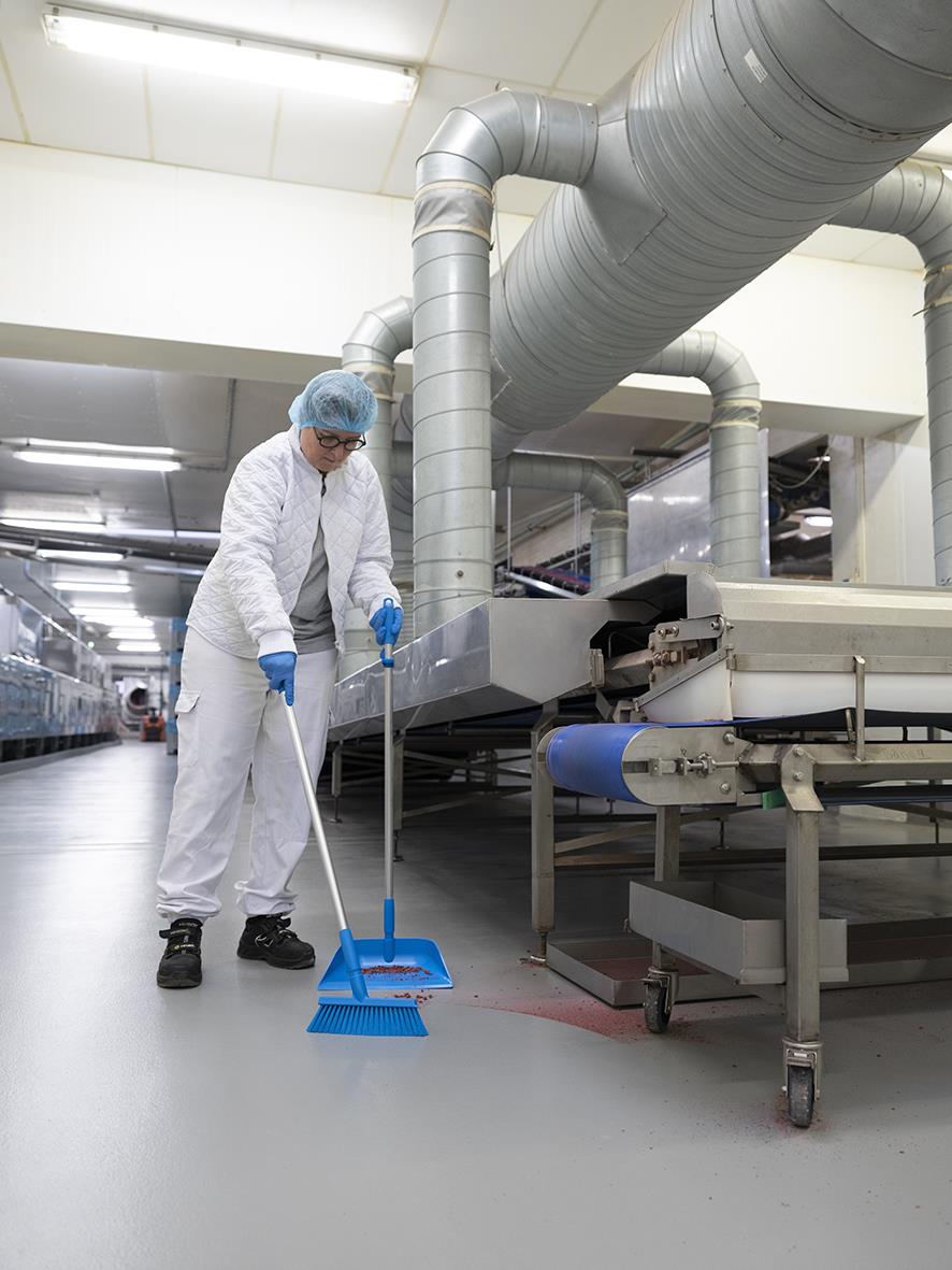 Person i skyddskläder och hårskydd sopar golvet i en industriell miljö med blå sopskyffel