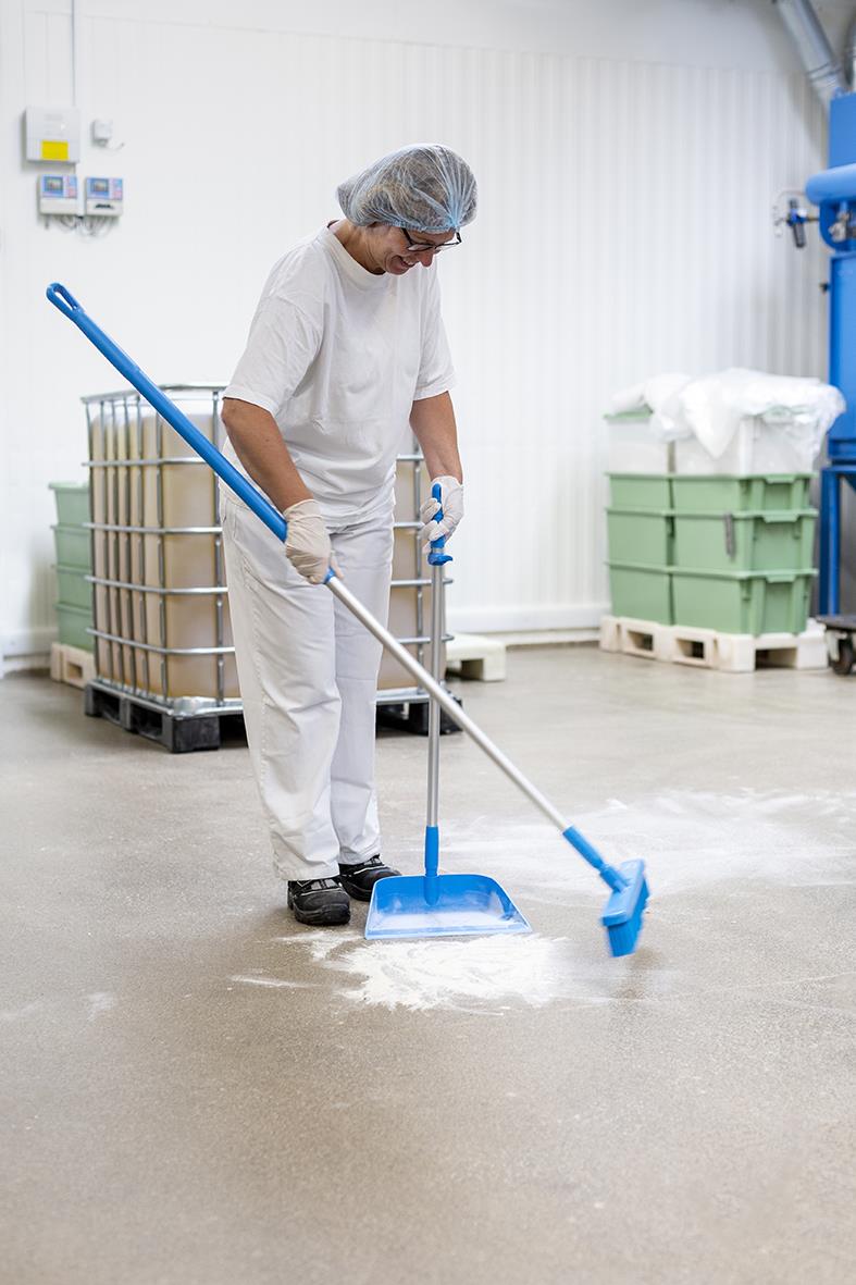 Person i skyddskläder använder sopskyffel Vikan stående blå 330mm för att sopa upp pulver på golvet i ett industriellt utrymme