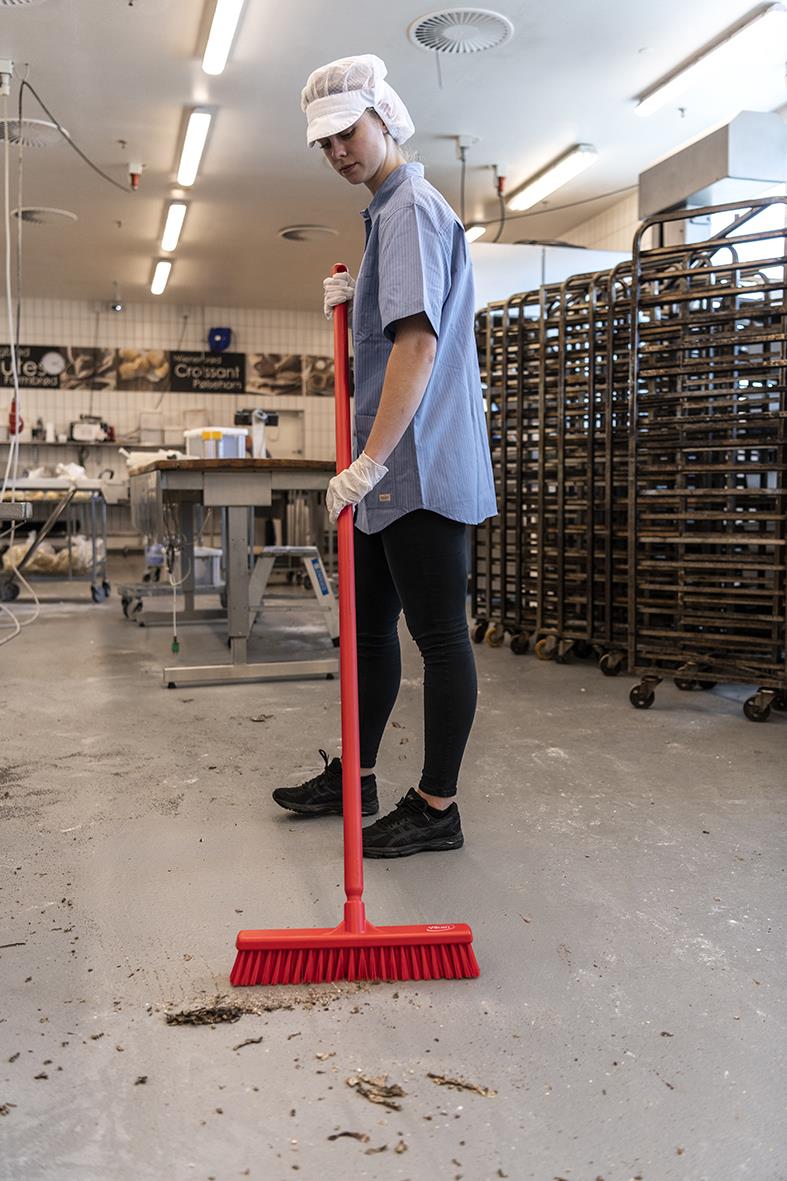 Person som sopar golvet med en röd golvsopborste i ett industriellt kök eller fabriksmiljö