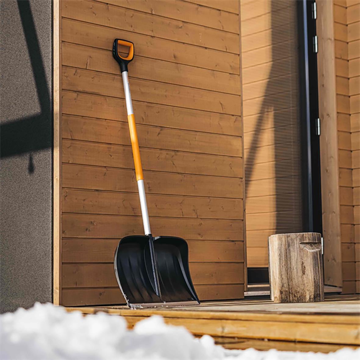 Snöskyffel från Fiskars X-Series med svart skopa och orange handtag stående mot en trävägg