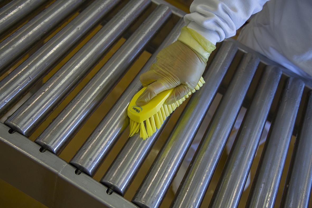 Person rengör metallrullar med en gul handborste i en industriell miljö