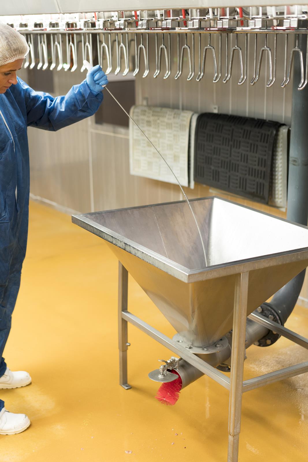 Person i blå arbetskläder och handskar håller ett flexibelt rostfritt stålhandtag över en industriell maskin i ett laboratorium eller fabrik