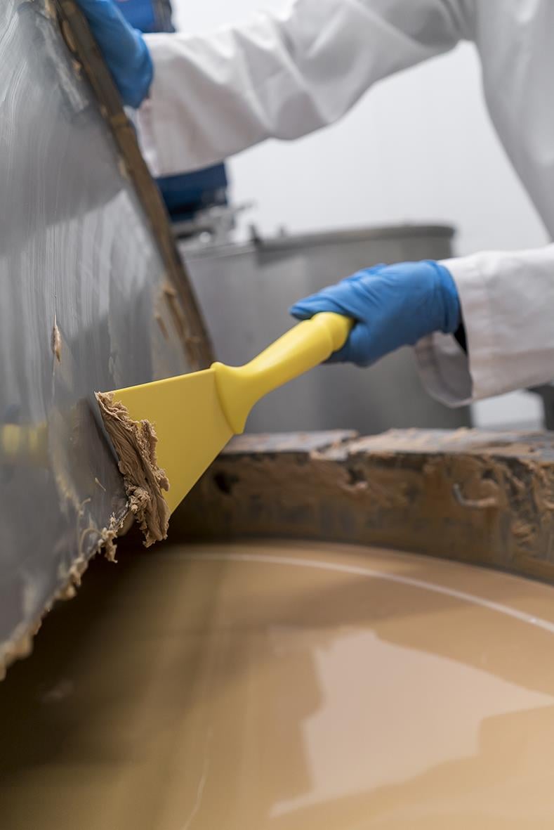 Person använder en gul spackelspade för att applicera material på en stor metallyta, med blå handskar och vit arbetsklädsel