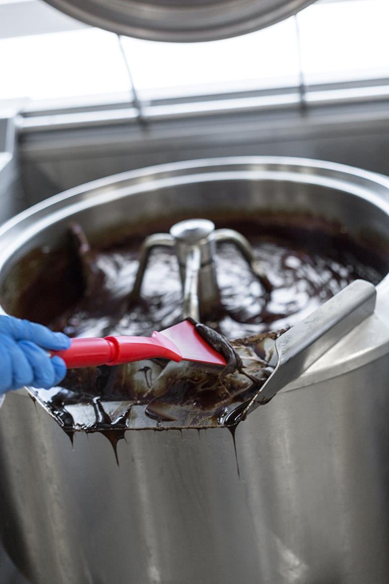 Person i blå handskar skrapar mörk vätska från en stor metallkittel med en röd skrapa