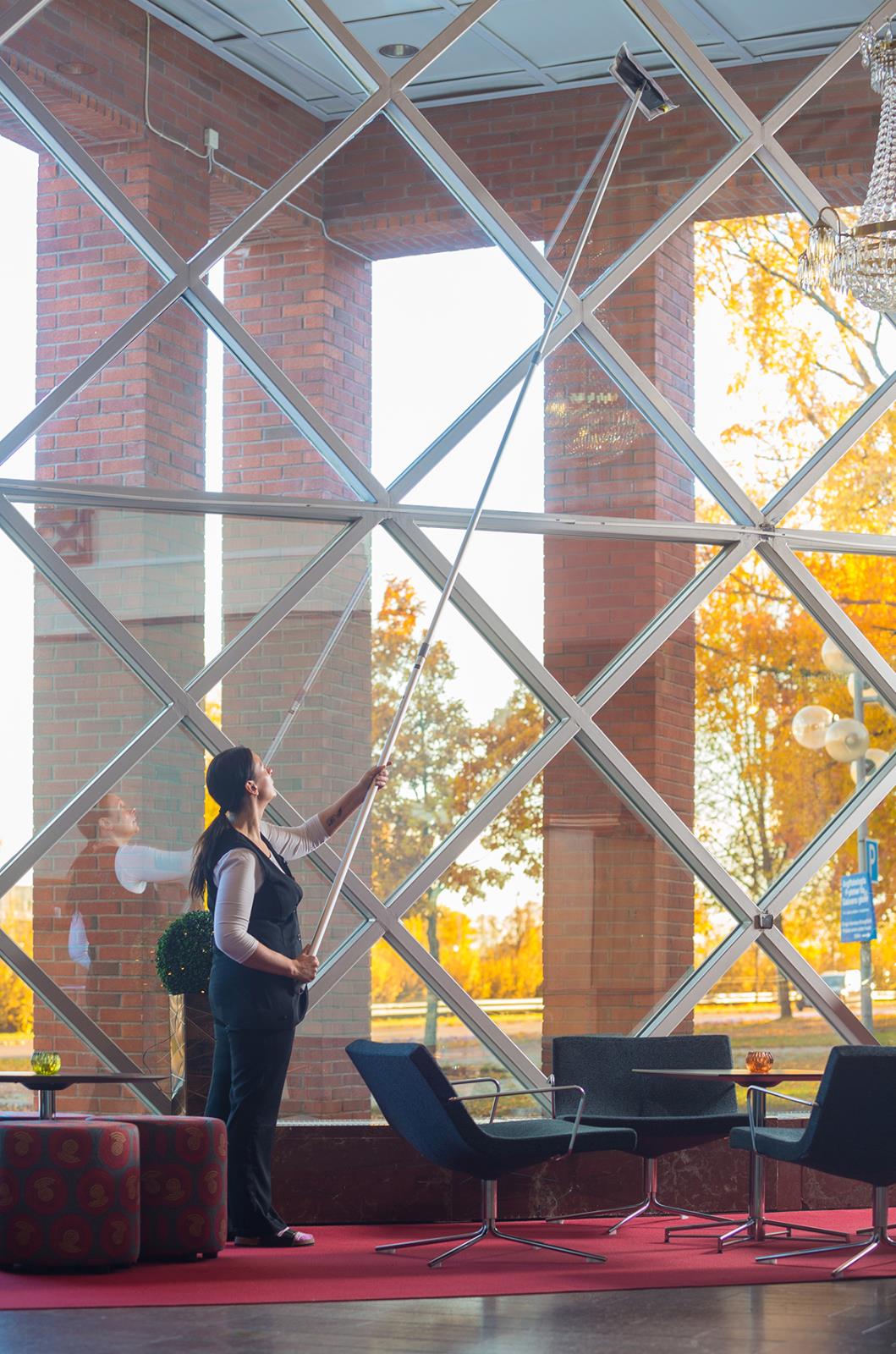 Kvinna rengör stor fönstervägg med teleskopskaft i ett modernt inomhusmiljö med stora glasfönster och utsikt utomhus i höstfärger