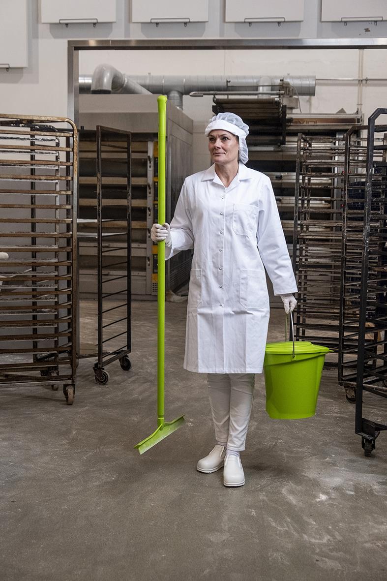Kvinna i arbetskläder med limegrön hink och moppskaft i ett industriellt kök eller bageri