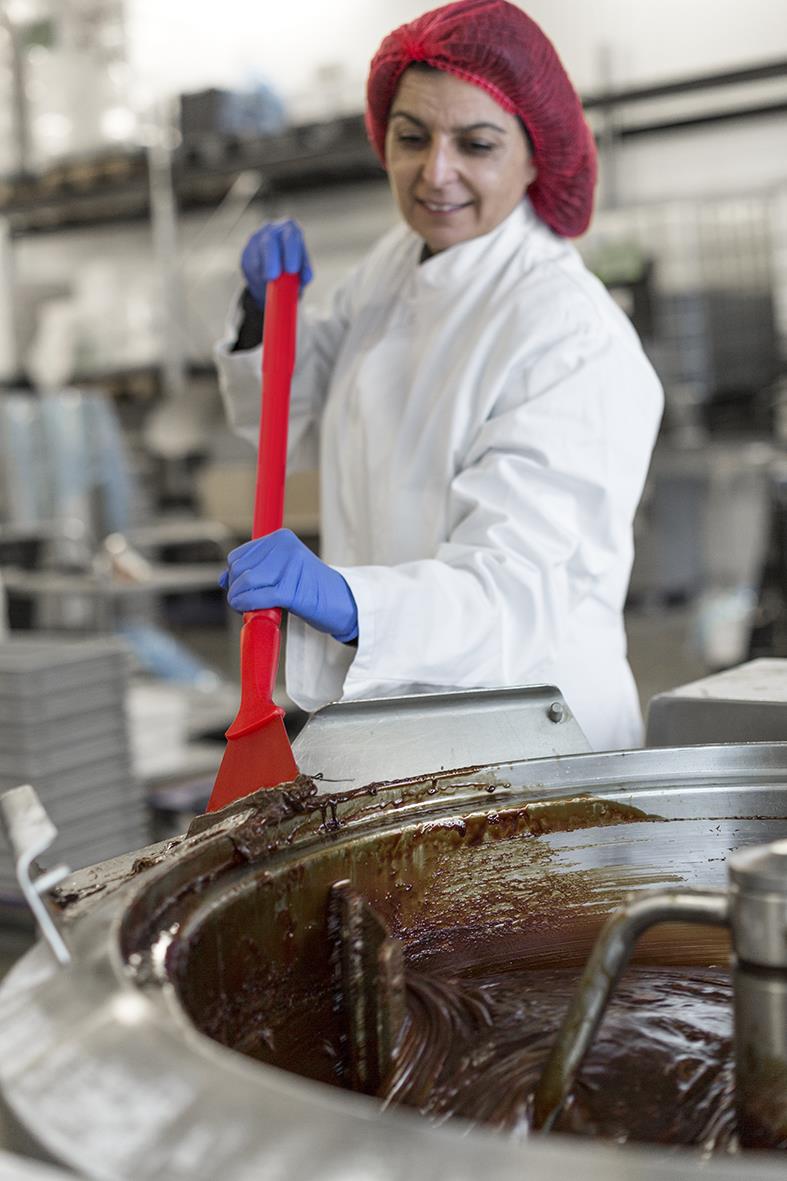 Person i skyddskläder och hårnät rör om i stor kastrull med choklad på ett kök eller bageri