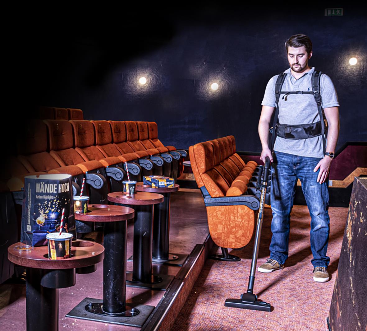 Man med ryggsäck som städar en biosalong med en bärbar dammsugare i mörkt rum med röda stolar