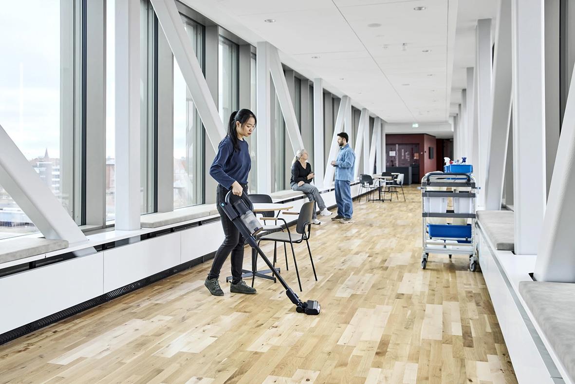 Person som städar med dammsugare i ett modernt kontorsutrymme med stora fönster och trägolv